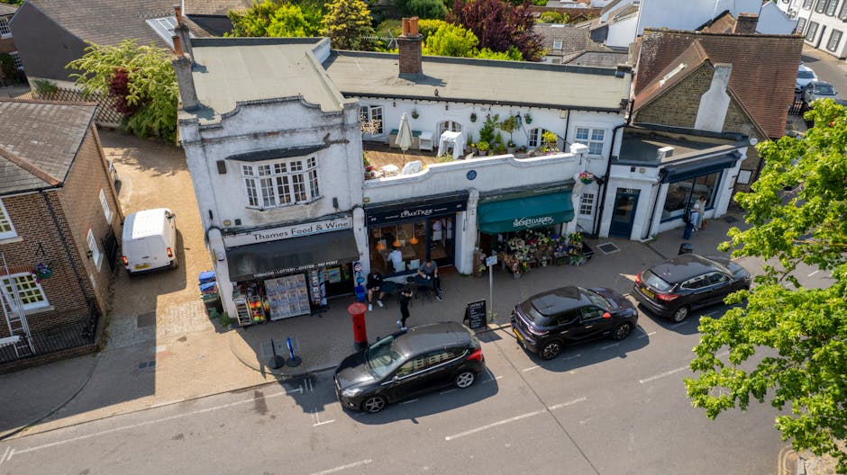 An aerial view of a quaint commercial street in Hampstead Village, featuring a row of small shops with white facades and large display windows, including a grocery store named Thames Food & Wine with 
